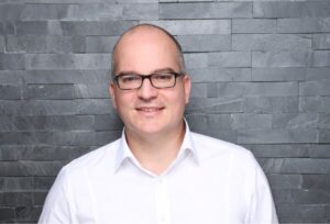 A person with glasses and a white shirt smiles friendly into the camera, standing in front of a wall of gray stones that are stacked horizontally. The photo looks professional and well-lit.