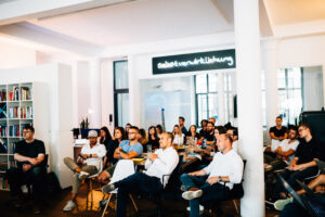 A group of people sit attentively on chairs and observe. There are bookshelves in the background and a sign on the wall that reads "Self-realization".