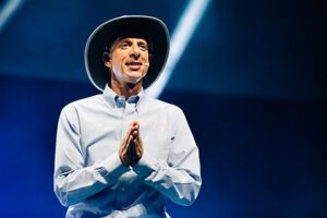 One person wears a wide hat and a light-colored shirt and holds her hands folded in front of her. She is smiling on a stage with a blue background and lighting effects.