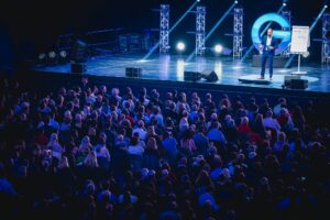 A man is speaking on a stage in front of a large audience. He gesticulates next to a flipchart. The stage is surrounded by blue lights and technical equipment. The audience listens attentively.