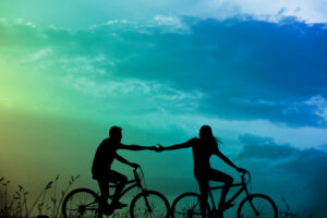 Two people are riding bicycles and holding hands. The background shows a sky with clouds and a color gradient from green to blue, while blades of grass are visible on the ground.