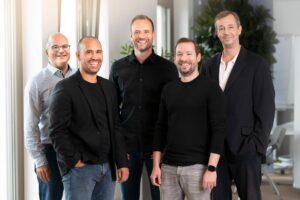 Five smiling men in casual clothes pose in a group in a bright, modern office with plants in the background. They stand upright and radiate a friendly atmosphere.
