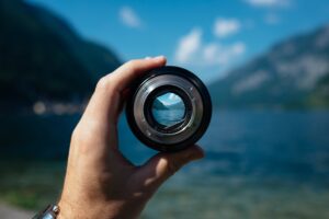 One hand holds a camera lens that focuses on a sharp landscape with water and mountains. The same landscape can be seen out of focus in the background.