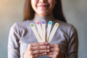 Wooden sticks with different smiley faces are held by one person in both hands. The person is wearing a gray sweater and smiling slightly in the background, in a neutral environment.