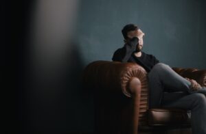 A person sits on a brown leather sofa and holds his head in a thoughtful gesture. The background is a dark green wall that creates a calm atmosphere.