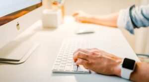 Ein Computer-Bildschirm auf einem Schreibtisch, eine Hand tippt auf einer wei&szlig;en Tastatur, w&auml;hrend eine andere Hand eine Maus bedient. Ein &Auml;rmel verdeckt teilweise den Arm.