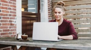 Eine Person sitzt konzentriert vor einem Laptop an einem Holztisch im Freien. Neben dem Laptop stehen eine Tasse Kaffee und eine Teekanne. Die Umgebung ist von Backsteinmauern und Holzw&auml;nden gepr&auml;gt.