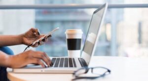 A person is typing on a laptop and holding a smartphone at the same time. Next to the computer is a paper cup with a lid on a white table. Glasses lie next to it. There are blurred windows in the background.