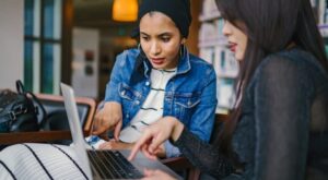 Zwei Personen sitzen im Innenbereich an einem Tisch und interagieren mit einem Laptop. Beide zeigen auf den Bildschirm. Im Hintergrund stehen B&uuml;cherregale. Die Szene wirkt studien- oder arbeitsbezogen.