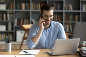 A person with glasses and a beard is smiling and talking on a smartphone. She is sitting in front of an open laptop at a desk with office supplies, surrounded by bookshelves.