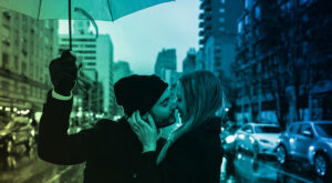 Man and woman kissing under an umbrella while standing in winter clothes on a rain-soaked, busy city street. Cars and tall buildings line the street.