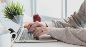 H&auml;nde tippen auf einer Laptop-Tastatur, begleitet von einer Kaffeetasse und einer Topfpflanze. Im Hintergrund steht ein roter Apfel auf einem wei&szlig;en Tisch, nahe einem Fenster.