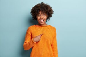 A person wears an orange sweater and points a finger at herself in surprise. She is standing in front of a light blue background. The facial expression and posture appear joyfully surprised.