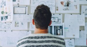Eine Person mit gestreiftem Pullover betrachtet aufmerksam eine Wand voller Notizen und Fotos. Die Umgebung wirkt kreativ und strategisch, m&ouml;glicherweise ein Arbeitsbereich f&uuml;r Planung oder Brainstorming.