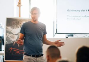 A man in a blue T-shirt gives a presentation to a seated audience. In the background a poster and a screen with text: "Winning the 1, [...] contract signing, sales launch."