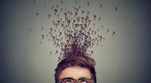 A man with glasses looks upwards while numerous question marks rise from his head. The background is grey, which emphasizes the question marks and symbolizes confusion or thoughts.