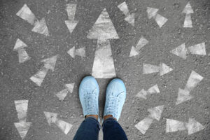 A pair of light blue sneakers stands on a gray asphalt floor. Surrounded by white arrows pointing in different directions, the image conveys the impression of decision-making and disorientation.