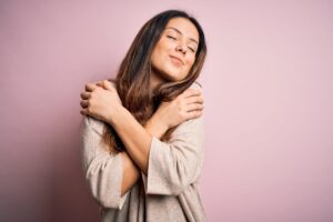 A person with long hair hugs herself and smiles with closed eyes, standing in front of a soft pink background, wearing a cozy beige top.