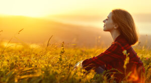 Eine Person sitzt entspannt, blickt l&auml;chelnd in den Sonnenuntergang und tr&auml;gt ein kariertes Hemd. Umgeben von einem goldenen, bl&uuml;henden Feld vermittelt die Szene Ruhe und Gl&uuml;ck.