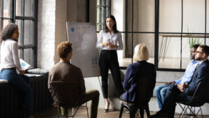 A woman stands in front of a flipchart, explains diagrams and speaks to five people sitting in a circle of chairs in a modern, industrial room. She concentrates on the presentation.