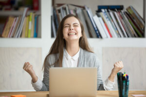 A young woman sits in front of a laptop with clenched fists and a broad smile, showing joy. Books can be seen on a shelf in the background, with pens in a holder next to them.