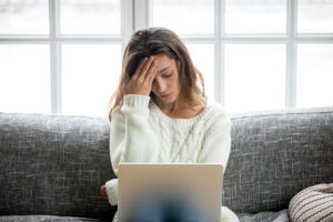Woman sitting on a gray sofa in front of a laptop, supporting her head with her hand, surrounded by large windows that let in bright daylight. She is wearing a white knitted sweater.