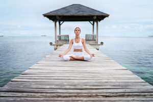 A person meditates in the lotus position on a wooden footbridge leading into the sea. A covered bench stands at the end of the jetty. The sky is cloudy, surrounded by calm water.