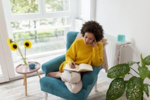 A person in a yellow sweater is sitting on a blue armchair, writing in a notebook. Next to her is a table with sunflowers and a cup. Large windows offer a view of the greenery.