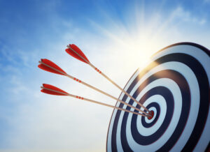 Three red arrows hit the center of a black and white target. The sun shines in the background. The sky is clear and blue, indicating an outdoor environment.