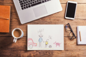 A child's picture shows people labeled DADDY and ME, a ball and a pet. Surrounded by a laptop, smartphone, glasses, notebook and coffee on a wooden table.