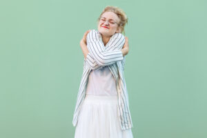 A person with glasses and a blonde pinned-up hairstyle hugs herself and smiles. She is wearing a striped jacket and a white skirt. The background is plain green.