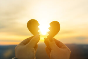 Two hands hold a wooden heart broken in half. The sunset in the background creates a warm, golden glow through the heart, which illuminates the sky.