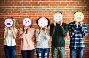 Five people hold colorful emoticon signs in front of their faces, showing different facial expressions, from sad to happy. They are standing in front of a brick wall and wearing casual clothes.