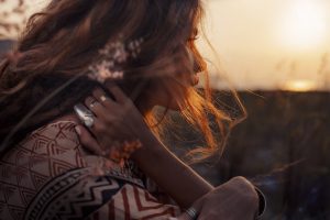 Eine Person sitzt in nachdenklicher Haltung bei Sonnenuntergang, tr&auml;gt einen gemusterten Pullover, h&auml;lt den Hals mit einer Hand. Die Szene wirkt ruhig und warm, umgeben von Natur.