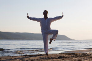A person in white clothing stands on one leg in a balancing yoga pose on the beach, while the calm sea and hills are visible in the background at sunset.