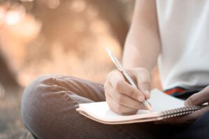 A person is sitting outdoors, writing in a notebook resting on his knee. The relaxed surroundings with a blurred background suggest a park or garden.