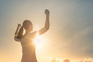 Person hebt die Arme in triumphaler Pose, w&auml;hrend sie von der aufgehenden oder untergehenden Sonne angestrahlt wird. Der Himmel ist klar, mit einigen Wolken am Horizont.