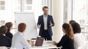 A man in a suit gives a presentation to a group of five people sitting at a table with laptops and coffee mugs; a flipchart shows drawn lines.