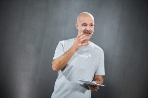 A bald man wears a gray T-shirt with the inscription Greator. He gestures with his right hand while holding papers in his left hand. The background is neutral gray.