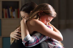 Two people embrace tightly and seem emotionally moved. The room is blurred in the background, with a bookshelf and warm light, creating a familiar, cozy atmosphere.