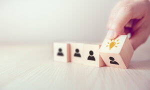 One hand grasps a wooden block with a symbol of a glowing light bulb, while two similar blocks with stylized figures are arranged on a table next to it.
