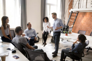 Six people sit and stand in a modern office while chatting. The room has large windows, natural lighting and brick walls. Some of the people are smiling as they listen attentively. There are two small plants on the table.