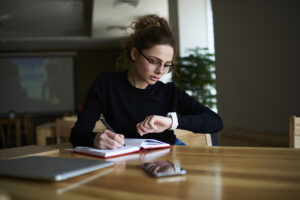 A person with glasses is sitting at a table, writing in a notebook and looking at their wristwatch. Tables and a projected image are blurred in the background.