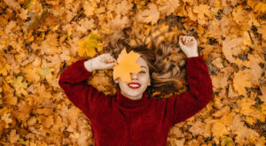A smiling woman lies on her back in a thick layer of orange autumn leaves. She is wearing a red sweater and holding a large leaf in front of one of her eyes.