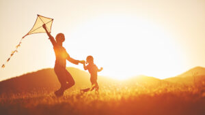 A woman and a child run across a meadow while flying a kite. The scene is at sunset, which creates a warm, golden lighting. Hills are visible in the background.