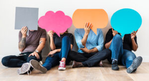 Vier Personen sitzen auf einem Holzboden, halten bunte, leere Sprechblasen vor ihre Gesichter. Die Sprechblasen sind in Grau, Rosa, Orange und Blau gehalten. Eine wei&szlig;e Wand bildet den Hintergrund.