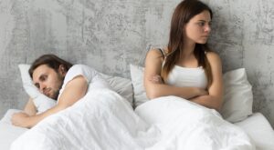 A man and a woman are lying in bed, the man is turned away and looks thoughtful, while the woman looks to the side with her arms crossed. The surroundings are a plain, gray room.