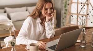 A woman sits smiling at a table, wearing headphones and looking at a laptop. Around her are vases of flowers and a cup, in a cozy, minimalistically furnished room.