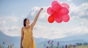 A person in a yellow dress holds several pink balloons, standing in a rural setting with mountains and flowering plants under a blue, cloudy sky.