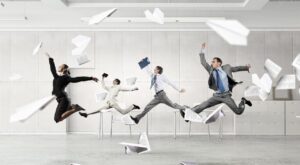 People in business attire skip joyfully through an office holding folders. In the background, sheets of paper remain floating in the air, above a sterile, modern office environment with desks and drawers.